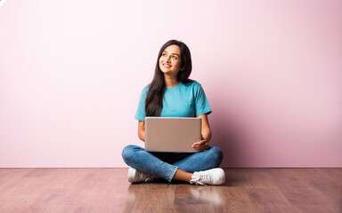 Indian asian girl using laptop while sitting against pink wall on wooden floor