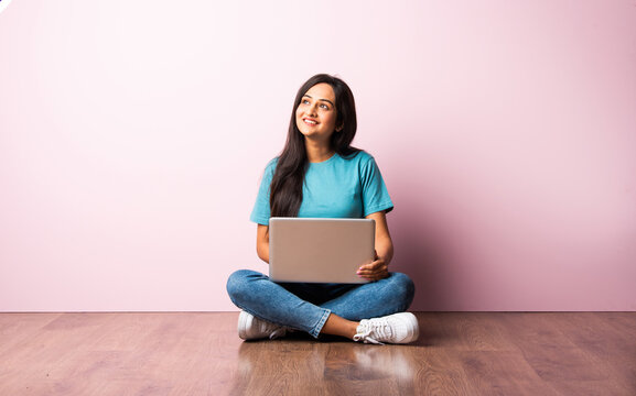 Indian Asian Girl Using Laptop While Sitting Against Pink Wall On Wooden Floor