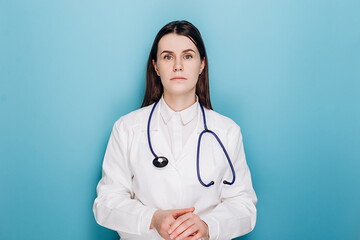 Anxious young woman doctor feels mental burnout at work, looking unhappy at camera, wears coat, stethoscope, isolated on blue background. Stressed frustrated physician upset about medical failure