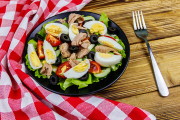Tasty tuna salad with lettuce, black olives, eggs and fresh vegetables on wooden table