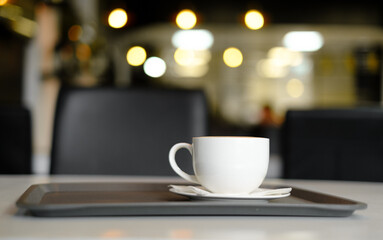 a white cup of coffee with a saucer on a black tray on a white table against the background of lights in a cafe with a black and white interior