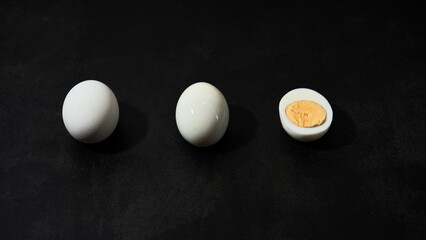 Minimalistic restaurant menu. Three chicken eggs lie on black kitchen table side view from below. Whole egg in white shell, peeled and cut in half so that you can see boiled yolk.