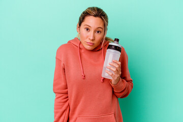 Young caucasian sport woman drinking water isolated on blue backgroundshrugs shoulders and open...