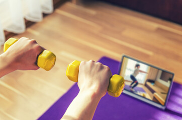 Young woman hands raising yellow dumbbells during workout sports at home, video exercises tutorials on tablet screen on floor mat in living room background, close-up view from back, sport concept