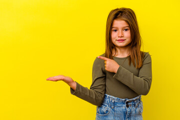 Little caucasian girl isolated on yellow background excited holding a copy space on palm.