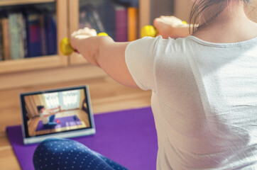 Young woman doing workout sports at home, raising dumbbells with straight arms, sitting on floor mat and watching video exercises tutorials on tablet screen in living room, close-up view from back