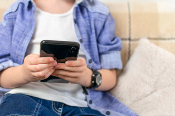 Young boy in casual wear sitting on sofa at home and using the phone. Child looks into a smartphone, types and sends message, playing a game at home. Technology, internet, leisure for kids and people