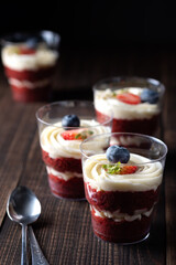 Trifle red velvet. Cake layered dessert in glass decorated with fresh berryes. In a small bowl are strawberry, blueberry, mint leave. Still life on a wooden table and dark backgroud with copy spase.