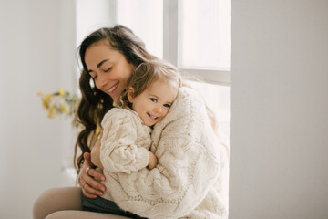 Naklejka premium Young mother hugging her little daughter, they sitting near window and smiling. 