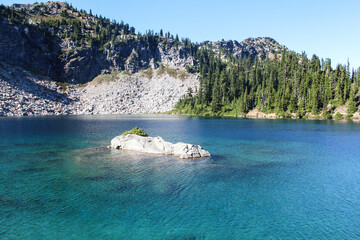 Naklejka premium beautiful crystal clear aquamarine lake with a big rock in the middle surrounded by mountains, pine trees and rocks