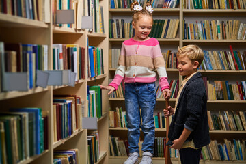 two school children help each other to get a book from shelf, stand talking, in library