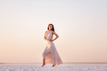 Young blonde woman in an evening airy pastel pink, powdery dress stands barefoot on white...