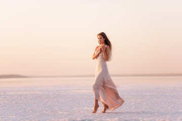 Young blonde woman in an evening airy pastel pink, powdery dress stands barefoot on white...