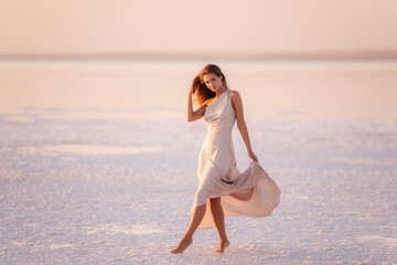 Young blonde woman in an evening airy pastel pink, powdery dress stands barefoot on white...