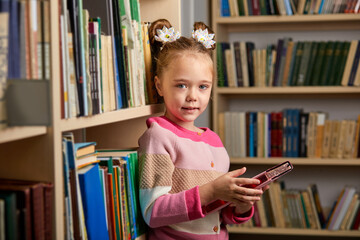 kid girl with ponytails stands in library after classes, enjoy being educated, get new information and knowledge
