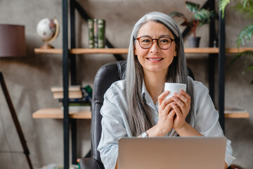 Emotional mature grey-haired woman watching movie on her laptop holding a cup of hot drink