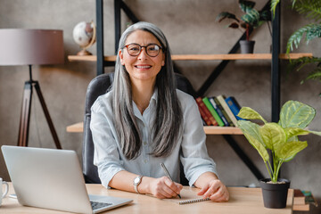 Cheerful middle-aged woman with grey hair working on laptop in office