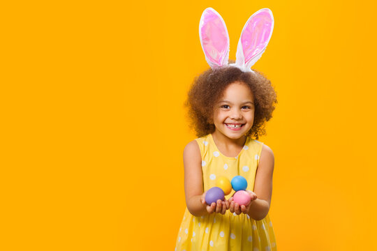 An african girl in sunny orange dress with rabbit ears on her head with painted eggs in her hands on a yellow background