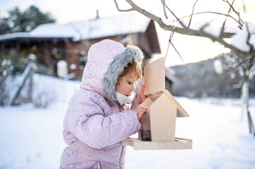 Small girl outdoors in winter garden, standing by wooden bird feeder.