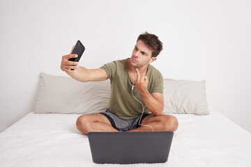 Angry young man taking a selfie with his cell phone sitting on his bed insulting with his finger....
