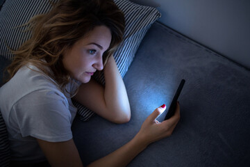 Young woman in bed holding a phone, tired and exhausted, blue light straining her eyes, messing up...