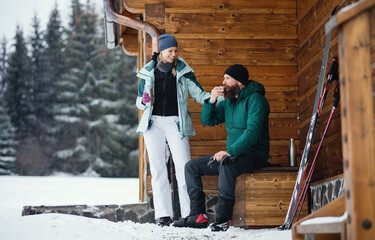 Mature couple drinking tea by wooden hut outdoors in winter nature, cross country skiing.