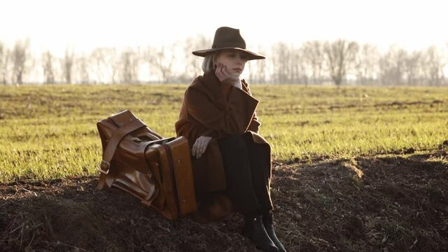 stylish girl in a hat with a suitcase sitting on the grass by the field in sunset