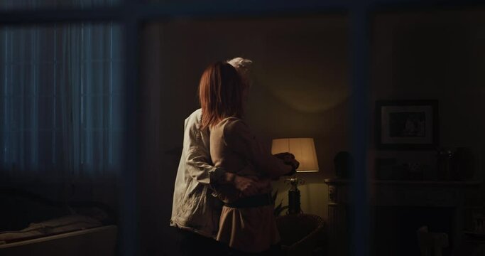Cinematic shot from window of young happy homosexual female gay and transgender man couple in love enjoying to embrace and kiss while dancing together and listening to the music at home at evening.