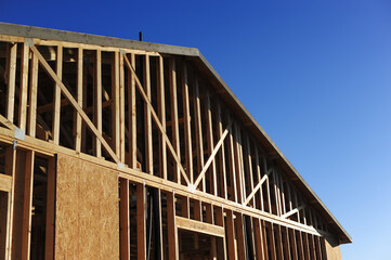 Obraz premium Close-up of gable-end framing with vertical studs, angled roof trusses, and partial sheathing on a residential building under construction against clear blue sky