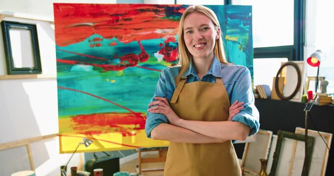 Portrait of young joyful Caucasian beautiful talented female artist in apron standing in studio workshop crossing hands looking at camera and smiling, pretty happy woman painter in room with big canva