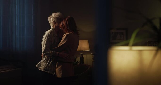 Cinematic shot from window of young happy homosexual female gay and transgender man couple in love enjoying to embrace and kiss while dancing together and listening to the music at home at evening.