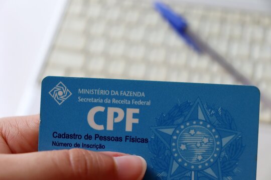 Bahia, Brasil - Fevereiro 3, 2021: Pessoa segurando o CPF - Cadastro de Pessoas Físicas (Physical Person Registration), com caneta e laptop ao fundo.