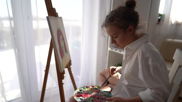Girl sits on a chair near the easel and holds a palette and a brush in her hands. Painting and hobby concept. Close up of a gentle girl in a white shirt painting a portrait on canvas