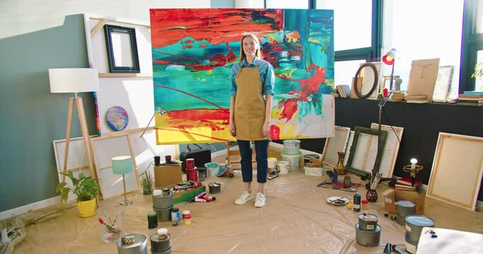 Caucasian young positive happy female painter in apron standing in own workshop with smile on face looking at camera and smiling, art gallery owner, joyful woman in studio near big canva, art concept