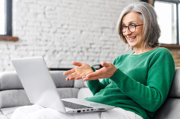 Positive senior mature woman in green jumper sitting on the couch, laughing and looking at the...