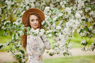 Young attractive girl stands among white blooming trees at the park. A woman wearing brown hat and...