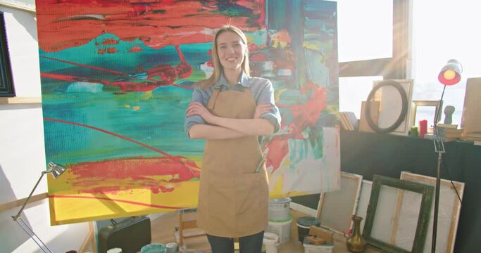 Camera approaching young happy Caucasian female talented painter in apron standing in own art studio crossing hands looking at camera and smiling in positive mood, canva in art gallery, workshop