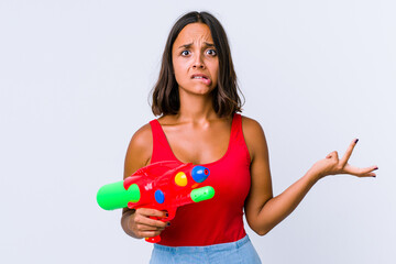 Young mixed race woman holding a water gun isolated confused and doubtful shrugging shoulders to hold a copy space.