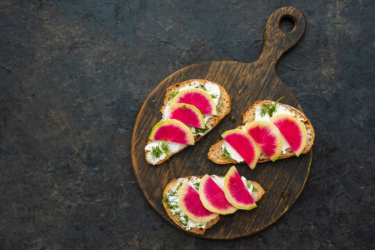 Light breakfast or snack, open sandwiches on whole grain bread with cream cheese, dill and watermelon radish slices on a wooden board against a dark concrete background. Sandwich recipes.