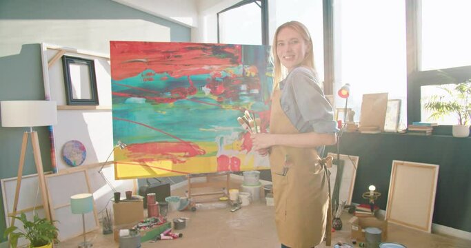 Rear of happy young Caucasian professional female artist in apron standing in art studio looking at finished painting on canva turning to camera holding paintbrushes and smiling to camera portrait