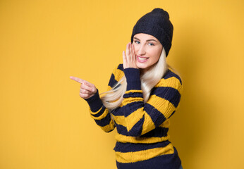 Woman holding hand near mouth and telling secret, model wearing woolen cap and sweater, isolated on yellow background. Whispering concept