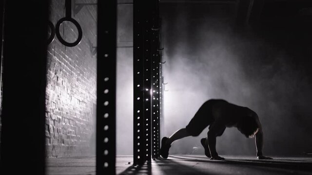 Wide shot of silhouette of muscular sporty man doing burpee while having intensive functional training in darkness in gym