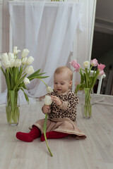 a little girl is sitting on the floor, dressed in a beige skirt, a jacket with a leopard pattern and red tights, holding a white tulip in her hands, looking at it, and next to her are two vases