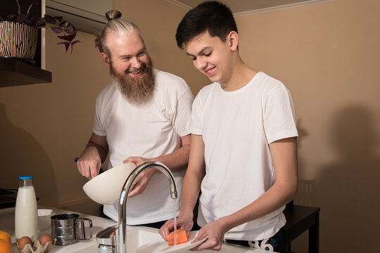 Happy Dad And Son Teenager On Kitchen. Smiling Father Cooking, Son Washing The Dishes. Concept Of Friendly Relationship Between Parents And Children Teens.