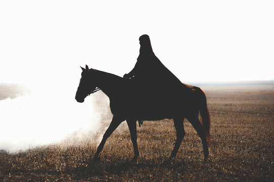 Beautiful Girl Riding A Horse Rider Walks Through The Smoke On The Background Field. Silhouette Of A Rider In A Cloak