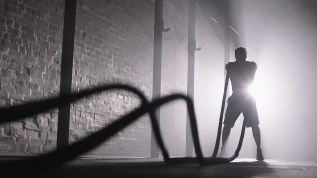 Wide shot of silhouette of muscular sporty man with naked torso doing exercises with battle ropes during crossfit training in dark gym