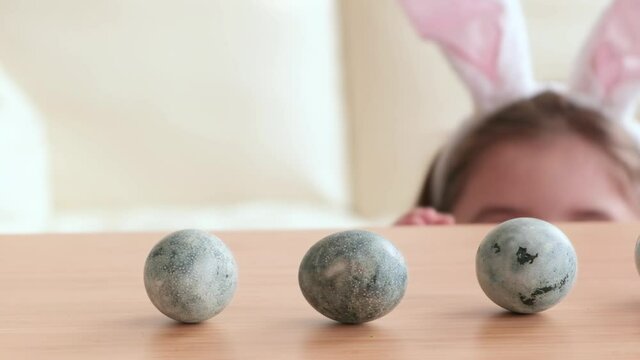 cute little girl wearing bunny ears playing egg hunt on Easter, hides under the table and picks up the egg. Adorable children celebrate Easter at home close up.