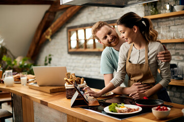 Happy couple talking while using touchpad and preparing food in the kitchen.