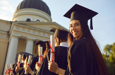 Portrait of a female graduate standing with a diploma in hand in a row with classmates and looking at the camera turning back. Education, graduation and people concept. © Studio Romantic