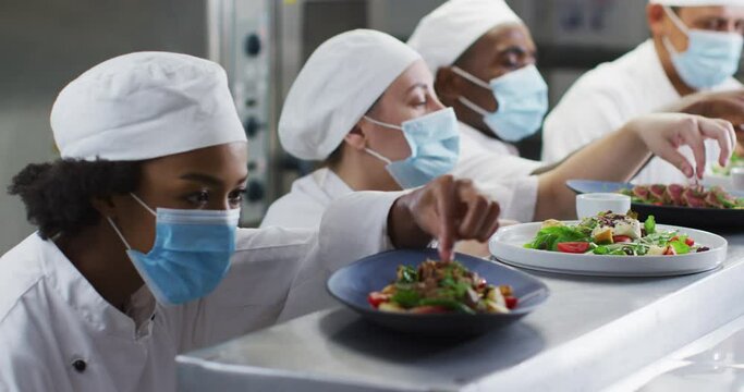 Diverse group of chefs wearing face masks garnishing dishes in restaurant kitchen